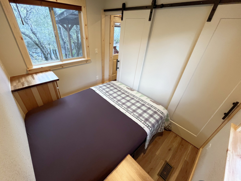 The master bedroom inside a Collins Lake cabin, with a queen bed, a window onto the oaks and a barn-door closet.