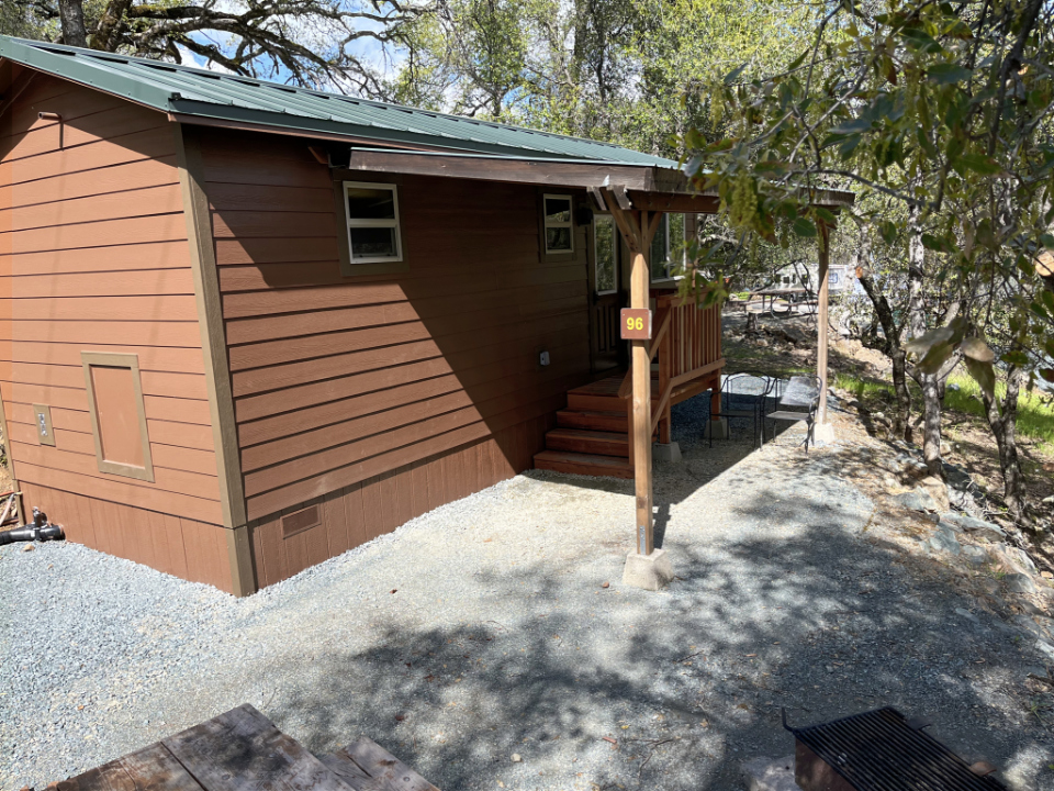 A brown timber cabin with a green metal roof and covered porch under oak trees at Collins Lake.