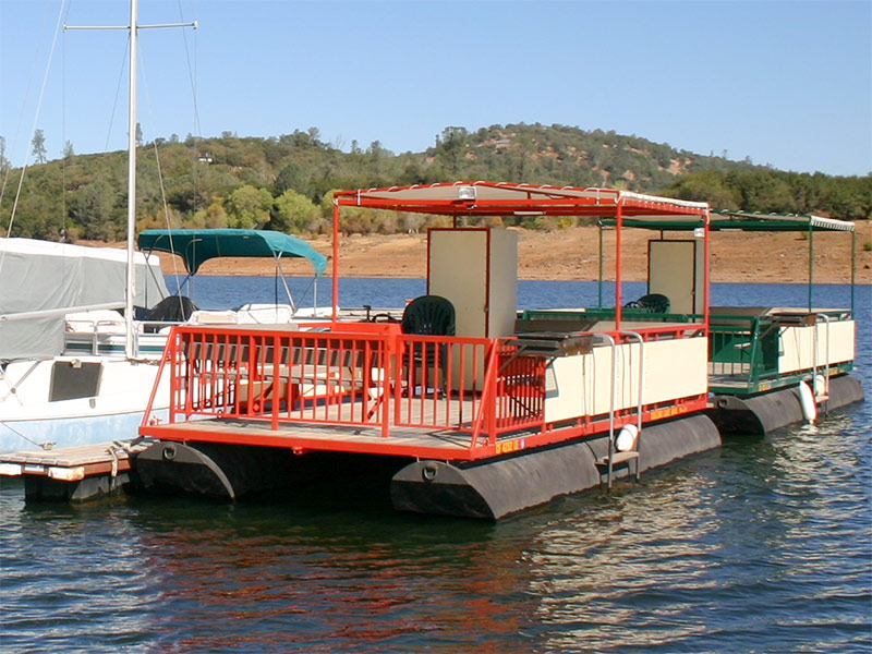 Two patio boats, one orange and one green, moored at the Collins Lake marina with the oak-covered shoreline behind.